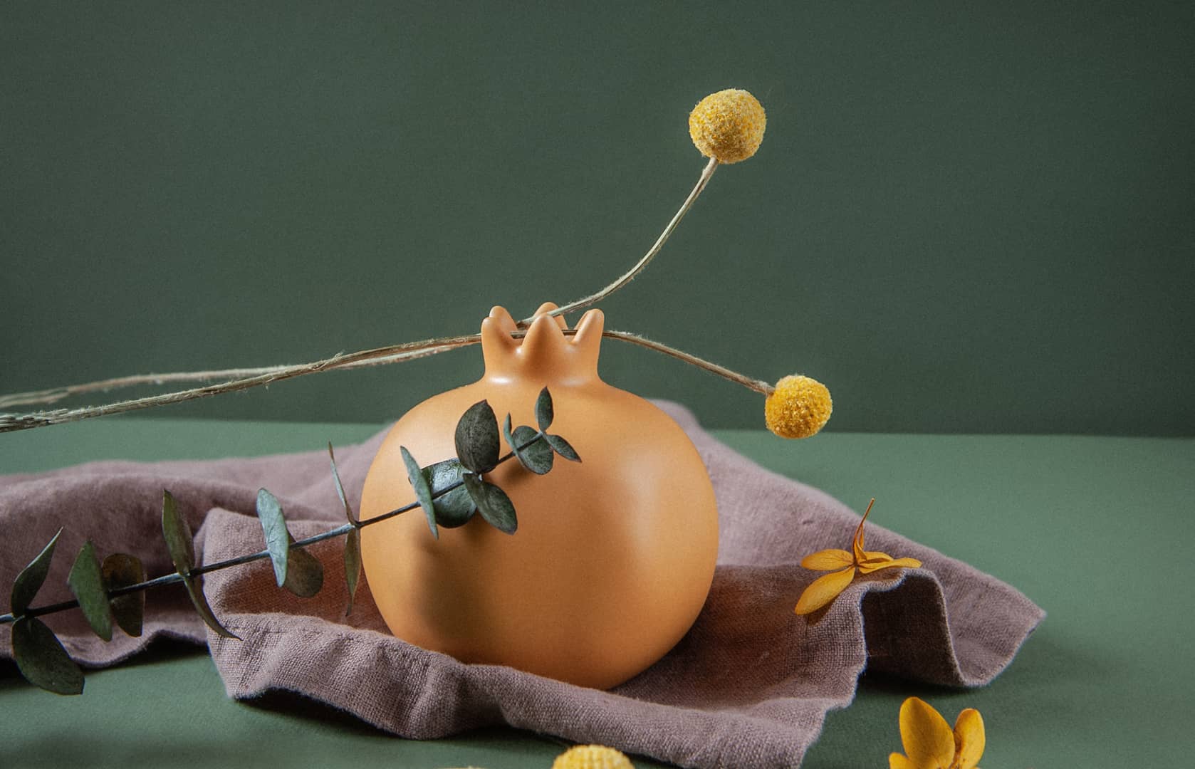 Vase en terre cuite orange orné de fleurs séchées sur un drap de lin prune, fond vert