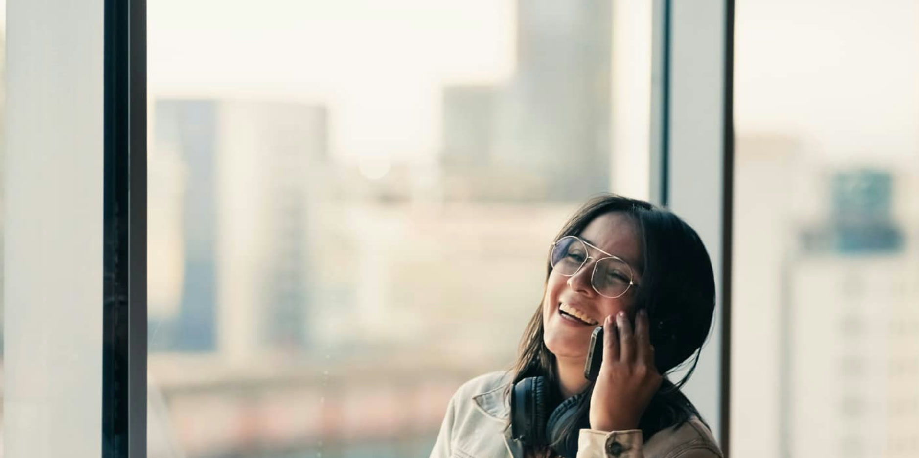 Jeune femme souriante au téléphone dans un bureau lumineux avec vue urbaine, illustration d'un investisseur particulier en train d'articuler ETF World et ETF thématiques